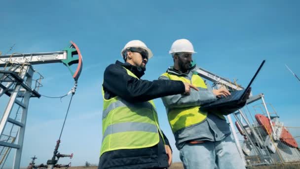 Deux hommes travaillent avec un ordinateur portable près de derricks de pétrole, plate-forme pétrolière . — Video