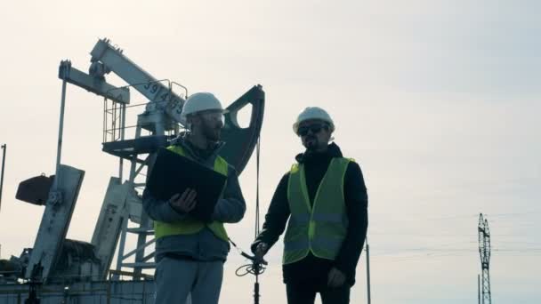 Engenheiros trabalham com um laptop perto de derricks de petróleo em um campo petrolífero. Conceito da indústria de extração de petróleo . — Vídeo de Stock