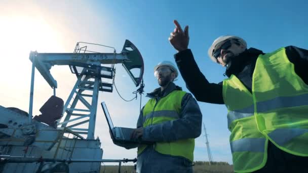 Mężczyzn z laptopem chodzić w pobliżu roboczych naftowej derrick. Kopalne paliwa, oleju przemysłu koncepcji. — Wideo stockowe