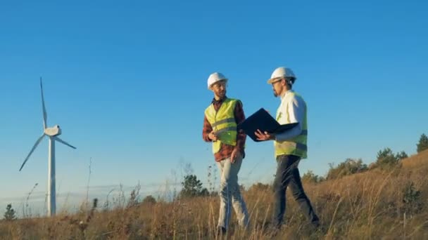 Een veld met twee energetics deskundigen lopen over het en een functionerende turbine-toren. Schone, eco-vriendelijke energieconcept. — Stockvideo