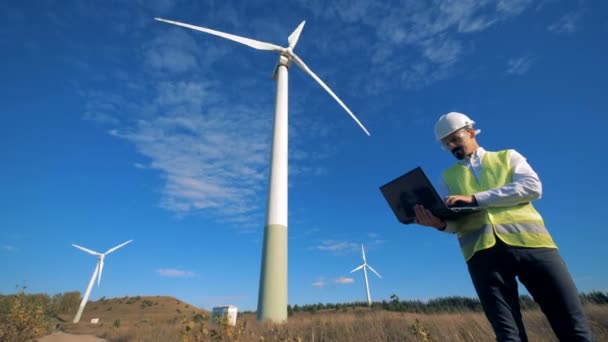 Sada větrných mlýnů se pracovník mužského působí jeho laptop vedle ní. Čisté, eko šetrné energetické koncepce. — Stock video