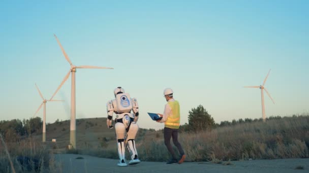 Un robot humain et un opérateur masculin marchent le long de l'ensemble des moulins à vent — Video