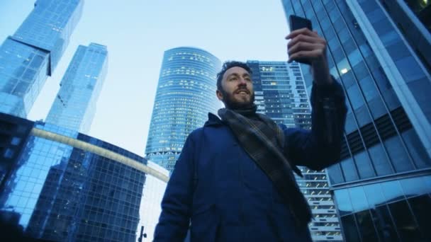 Um homem fazendo vídeo em seu smartphone enquanto está de pé na frente de edifícios de negócios — Vídeo de Stock