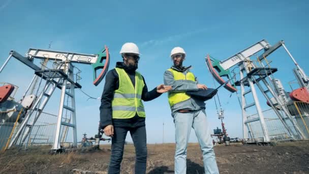 Zwei männliche Arbeiter unterhalten sich vor funktionierenden Ölförderanlagen. Energie, Öl, Gas, Treibstoffpumpe. — Stockvideo