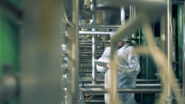 Работник работает над планшетом на заводе, окруженном трубопроводами — стоковое видео