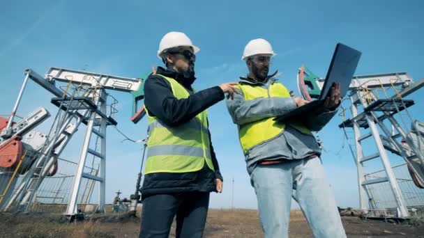 Два чоловіки працюють біля нафтових веж. Люди стоять біля нафтових викидів, працюють з ноутбуком . — стокове відео