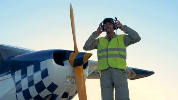 Piloto perto de avião pequeno, de pé com fones de ouvido em uma pista . — Vídeo de Stock