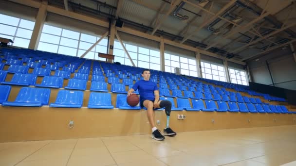 Ένας άνθρωπος με ένα βιονικό πόδι κάθεται με μια μπάλα του μπάσκετ σε ένα ευρύχωρο γυμναστήριο — Αρχείο Βίντεο