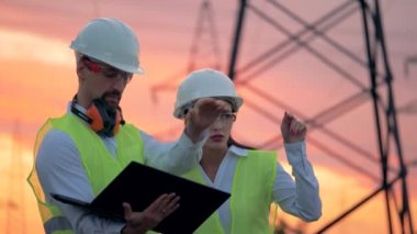 İki mühendis yenilenebilir enerji geliştirme üzerinde çalışan elektrik iletim hatları önünde konuşuyorsun.