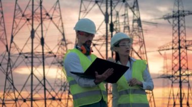 Elektrik iletim hatları ve iki teknisyen onlara yakın bir dizüstü bilgisayar ile çalışma. Yeşil enerji istasyonu inşa planı üzerinde ortak çalışma arkadaşları.