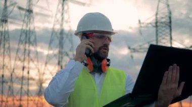 Bir adam iş giysisi içinde Etl kuleleri önünde bir telefonda konuşuyor