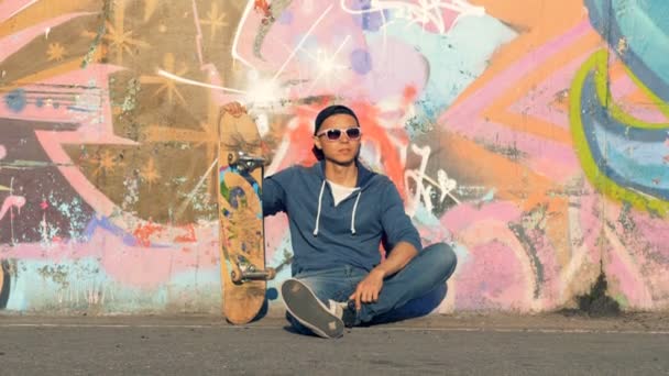 Adolescente masculino está sentado perto de uma parede de grafite com seu skate — Vídeo de Stock