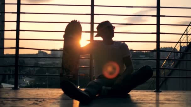 Stadtlandschaft bei Sonnenuntergang mit einem Jungen, der mit seinem Skateboard auf einer Treppe sitzt — Stockvideo