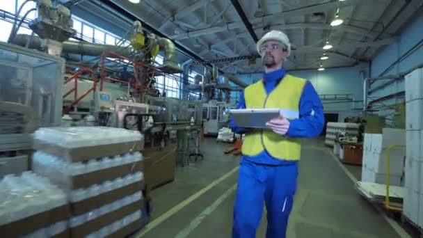 Male worker walks in a facility, typing on a tablet, factory equipment. — Stock Video