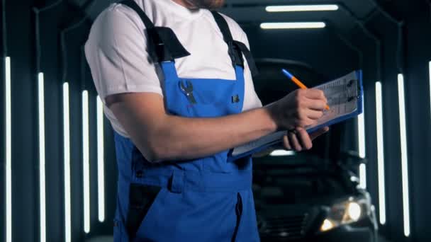 Ο άντρας μηχανικός γράφει μια αναφορά αφού επιθεωρήσει ένα αυτοκίνητο. Ιδέα υπηρεσίας αυτοκινήτου. — Αρχείο Βίντεο
