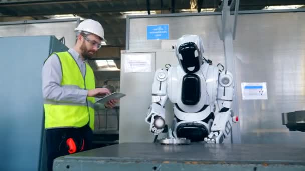 Un hombre escribe en una tableta mientras que un cyborg trabaja con un detalle de fábrica . — Vídeos de Stock