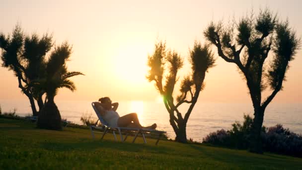 Uma mulher deitada em uma espreguiçadeira em um fundo marinho durante o nascer do sol. Conceito de férias de verão . — Vídeo de Stock