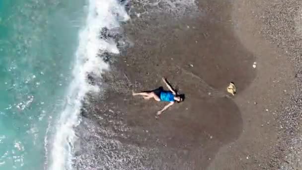 En flicka ligger på en strand i vatten medan vågorna slår mot stranden. Sommarsemester koncept. — Stockvideo