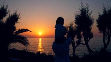 Omuzlarında bir adam ve bir çocuk denize el sallıyor.