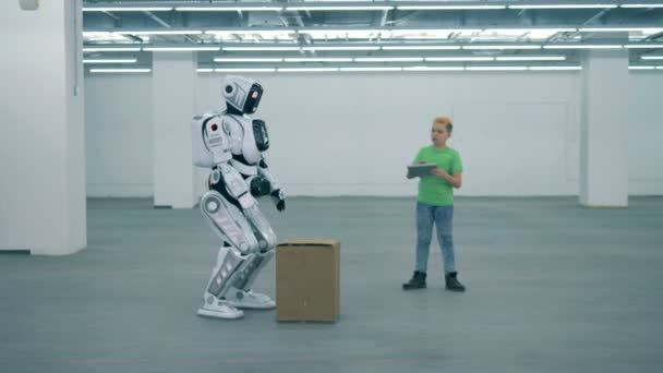 Bílý kyborg zvedne krabici, zatímco chlapec pracuje s tabletem, zblízka. — Stock video