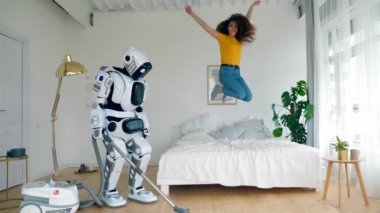 Bir robot zemin temizlerken mutlu kız yatakta atlar. Robot, cyborg ve insan konsepti.