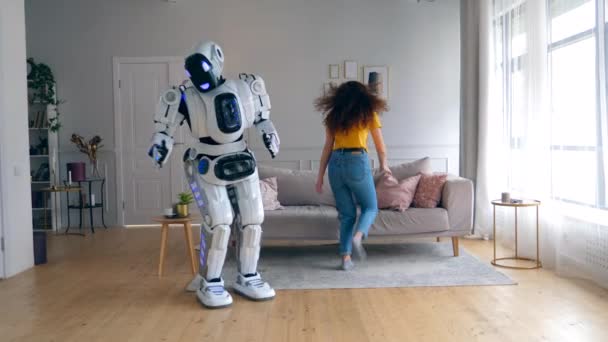 Chica alegre salta en un sofá mientras un droide baila. Robot, cyborg y concepto humano . — Vídeo de stock