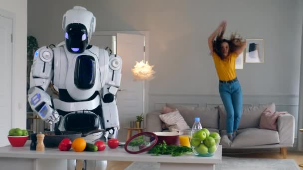 Chica alegre salta en un sofá mientras un robot cocina la cena . — Vídeos de Stock