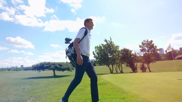 Ένας άντρας γκολφέρ περπατάει σε ένα χωράφι, κουβαλώντας σακούλα με εξοπλισμό. Παίκτης γκολφ σε γήπεδο γκολφ. — Αρχείο Βίντεο
