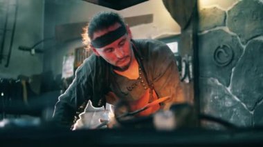 Forge işçisi metal bıçağı ısıtır, maşalarla tutar.