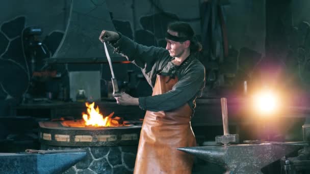 Professionele smid onderzoekt een metalen mes, staande op een smederij. — Stockvideo