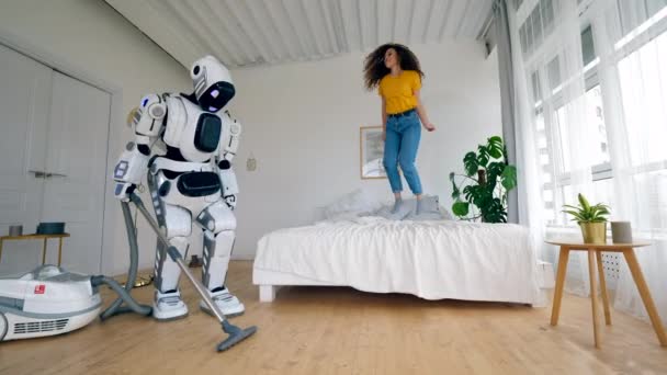 Jonge vrouw springt op bed terwijl een robot reinigt vloer. — Stockvideo