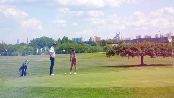 Uma menina bate uma bola em um campo de golfe enquanto um homem está assistindo . — Vídeo de Stock