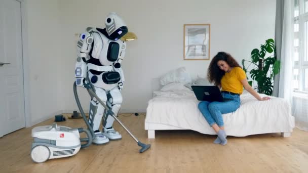 Een dame zit met een laptop en een robot is het schoonmaken — Stockvideo