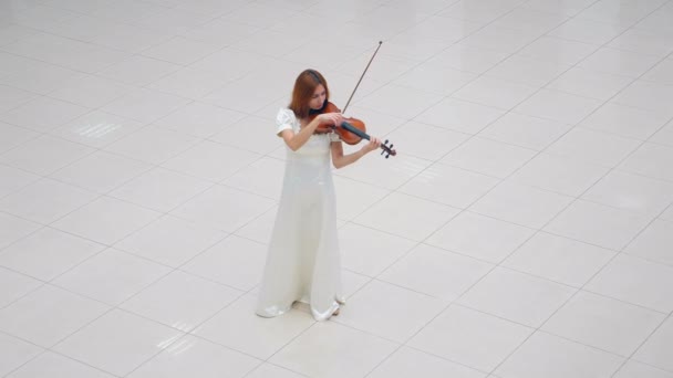 Жінка в білій сукні грає на скрипці, стоячи на білій плитці — стокове відео