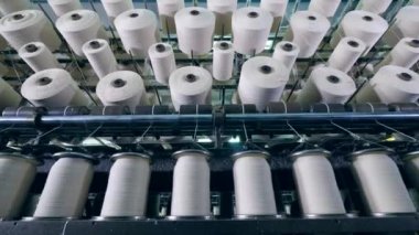 Fabrika makineleri bobinleri döndürüyor, beyaz iplikleri kıvrılıyor.