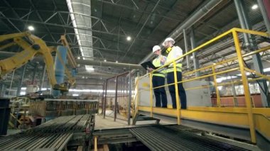 İki teknisyen modern fabrikada robot kolunu inceliyor.