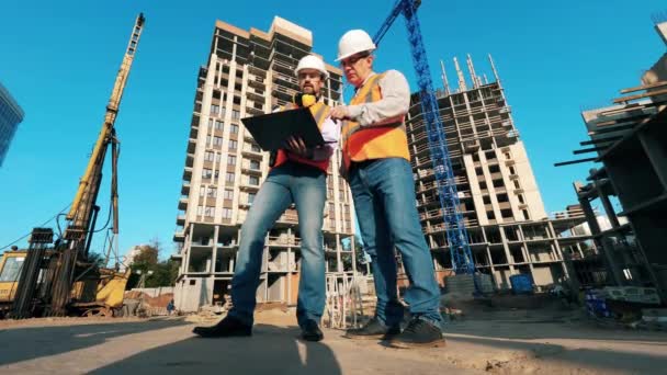 Два инженера разговаривают рядом с высотными зданиями, строящимися на заднем плане . — стоковое видео