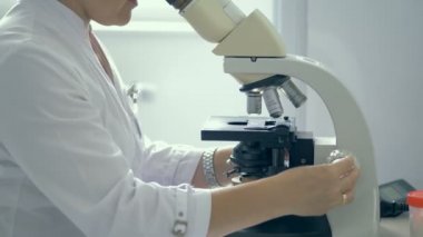 Kadın laboratuvar işçisi bir mikroskop u izliyor.