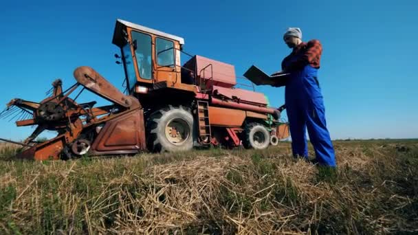 Farmer stands on a field with a laptop, checking tractor. — Stock Video