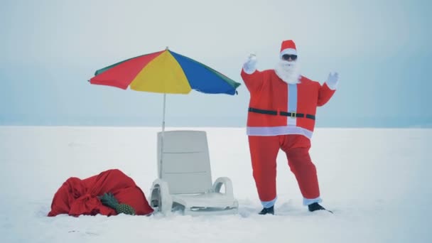 デッキチェアとビーチの傘の隣の雪の中でサンタクロースを踊る — ストック動画