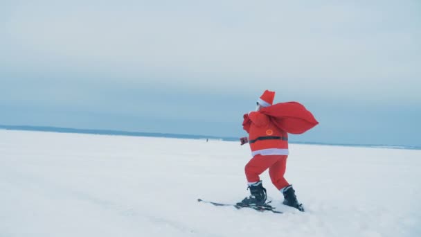 サンタクロースは雪の上にプレゼントの袋とスキーです。 — ストック動画