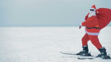 Karlı arazide Noel Baba kayak yapıyor ve ellerini sallıyor.