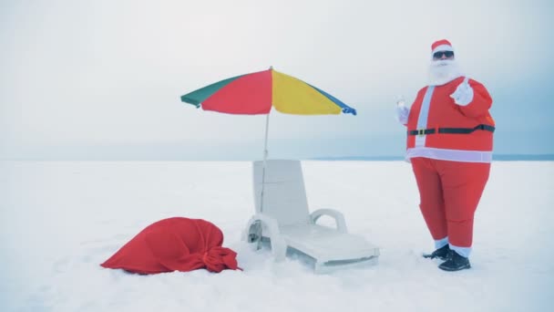 サンタクロースはデッキチェアと傘の近くで飲んで踊っています。 — ストック動画