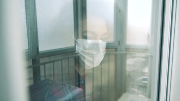 Uma mulher usa máscara médica e olha através da janela dentro de casa durante a pandemia . — Vídeo de Stock