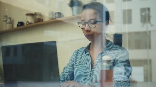 Eine Frau, die einen Laptop bedient, wird durch ein Café-Fenster gesehen — Stockvideo
