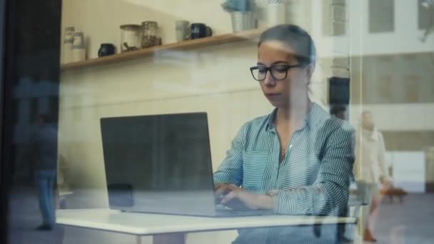 Una señora con gafas trabajando con una computadora vista por la ventana — Vídeos de Stock