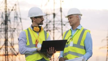 İki güçlü adam dışarıda elektrik kulelerinin yanında konuşuyor.