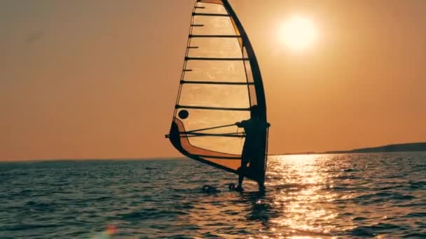 Um homem está atravessando as águas do pôr do sol em uma prancha de windsurf — Vídeo de Stock