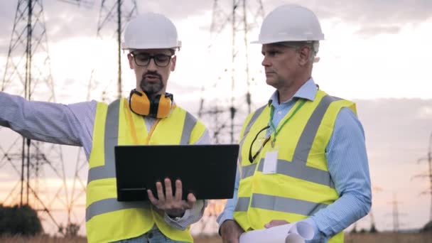 Dois homens trabalham com laptop em campo com linhas de energia. — Vídeo de Stock