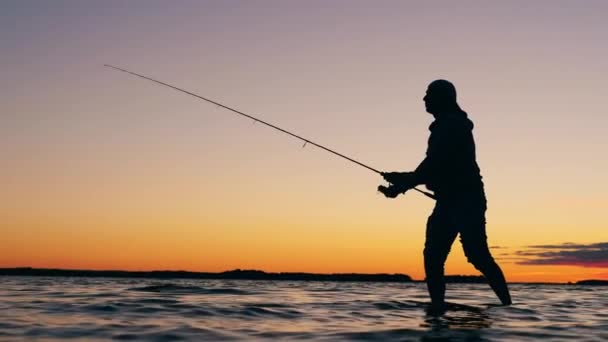 Ένας άντρας ψαρεύει σε μια λίμνη με καλάμι ψαρέματος το ηλιοβασίλεμα. — Αρχείο Βίντεο